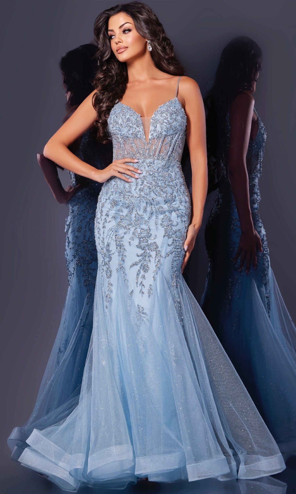 A woman poses in front of a grey background, her reflection visible, wearing the Jovani 43845 Long Prom Dress—a fitted light blue gown with floral sequin embroidery and a tulle skirt.